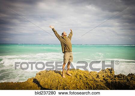Stock Image - Caucasian man standing on rocks at ocean with arms raised. Fotosearch