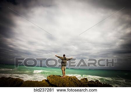 Stock Image - Caucasian man standing on rocks at ocean with arms outstretched. Fotosearch