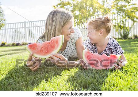 Stock Photo - Caucasian mother and daughter laying in grass eating watermelon. Fotosearch