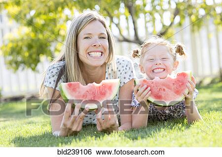 Stock Photograph - Caucasian mother and daughter laying in grass eating watermelon. Fotosearch