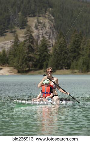 Stock Image - Caucasian mother and son sitting on paddleboard on lake. Fotosearch