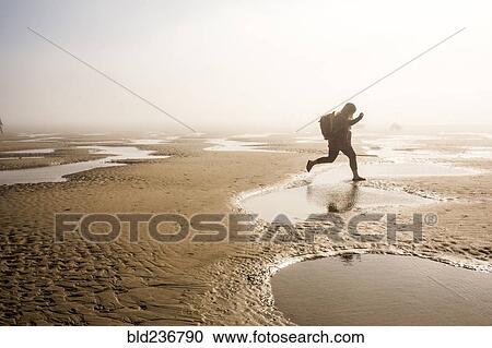 Stock Image - Caucasian mother carrying daughter jumping over puddle at beach. Fotosearch