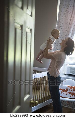 Stock Image - Caucasian mother holding baby son face to face in bedroom. Fotosearch