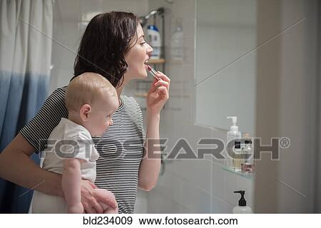 Stock Photo - Caucasian mother holding baby son while applying lipstick in mirror. Fotosearch