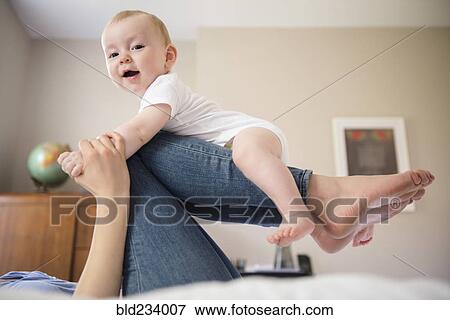 Stock Photo - Caucasian mother laying on bed balancing baby son on legs. Fotosearch