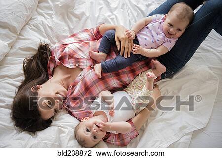 Stock Image - Caucasian mother laying on bed with twin baby daughters. Fotosearch