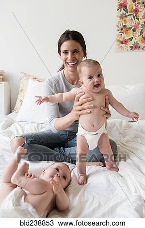 Stock Image - Caucasian mother playing with twin baby daughters on bed. Fotosearch