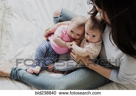 Stock Image - Caucasian mother sitting on bed holding twin baby daughters. Fotosearch