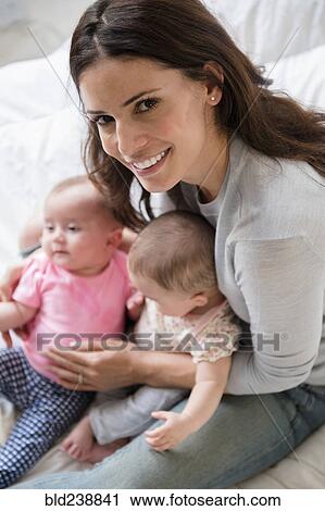 Stock Image - Caucasian mother sitting on bed holding twin baby daughters. Fotosearch