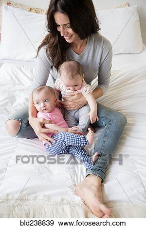 Stock Photo - Caucasian mother sitting on bed holding twin baby daughters. Fotosearch