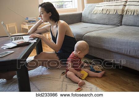 Stock Photograph - Caucasian mother sitting on floor using laptop near baby son. Fotosearch