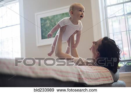 Stock Photo - Caucasian mother sitting on sofa lifting baby son. Fotosearch