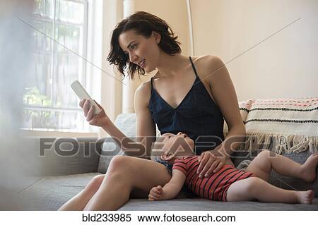 Stock Photography - Caucasian mother sitting on sofa with baby son texting on cell phone. Fotosearch