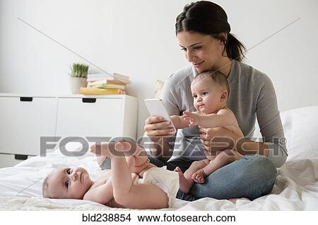 Caucasian mother with twin baby daughters on bed texting on cell phone View Large Photo Image Picture - Caucasian mother with twin baby daughters on bed texting on cell phone. Fotosearch
