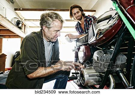 Stock Photo - Caucasian son watching father repairing motorcycle in garage. Fotosearch