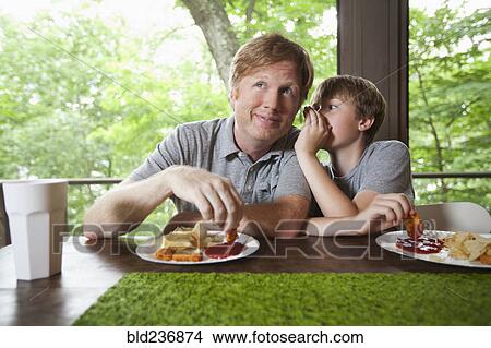 Picture - Caucasian son whispering to father and dipping food in ketchup. Fotosearch