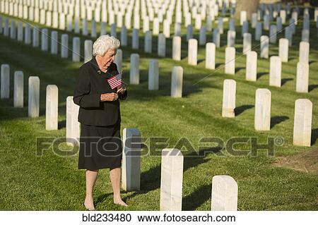 Stock Image - Caucasian widow holding American flag at cemetery gravestone. Fotosearch