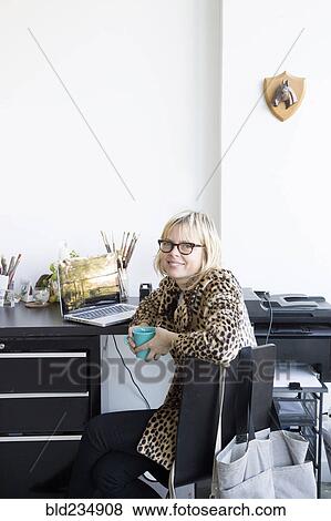 Stock Photo - Caucasian woman drinking coffee at desk with laptop. Fotosearch