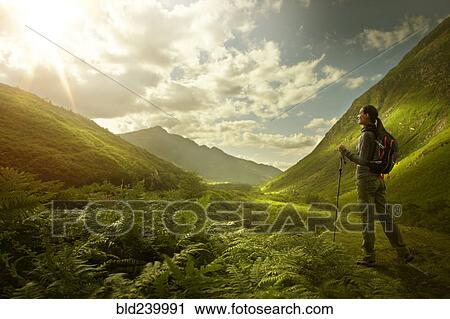 Stock Image - Caucasian woman hiking in green valley. Fotosearch