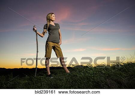 Stock Image - Caucasian woman holding walking stick at sunset. Fotosearch