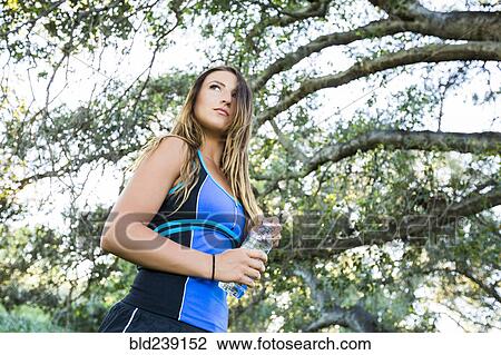 Stock Image - Caucasian woman holding water bottle under tree branches. Fotosearch