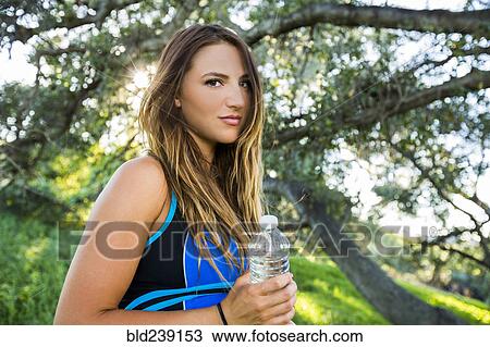 Stock Image - Caucasian woman holding water bottle near tree. Fotosearch