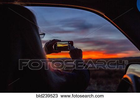 Stock Image - Caucasian woman in car photographing sunset with cell phone. Fotosearch