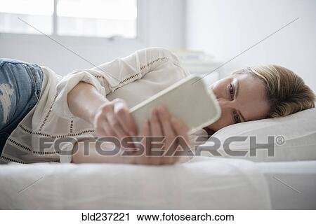 Stock Image - Caucasian woman laying on bed texting on cell phone. Fotosearch