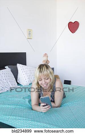 Stock Photo - Caucasian woman laying on bed texting on cell phone. Fotosearch