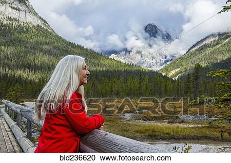 Stock Image - Caucasian woman leaning on wooden railing admiring mountain. Fotosearch