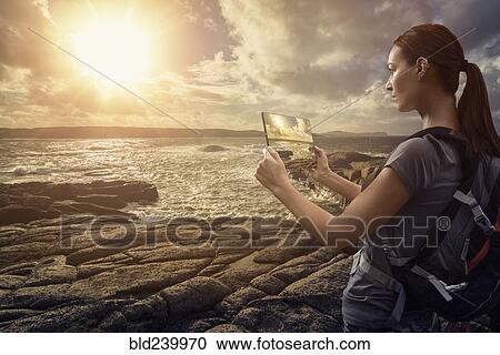 Stock Image - Caucasian woman photographing ocean sunset with digital tablet. Fotosearch