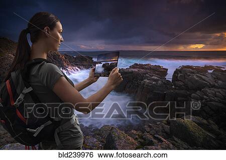 Stock Photograph - Caucasian woman photographing ocean sunset with digital tablet. Fotosearch