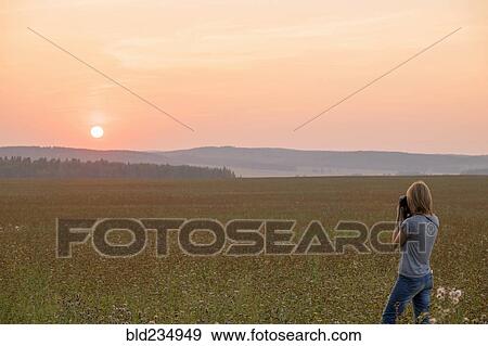 Stock Photo - Caucasian woman photographing sunset in field. Fotosearch