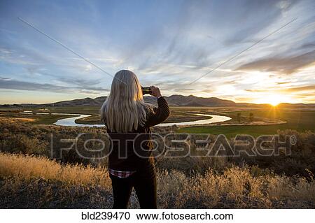 Stock Image - Caucasian woman photographing sunset over winding river. Fotosearch