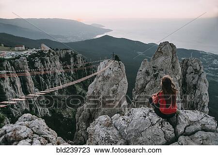 Stock Image - Caucasian woman sitting on mountain near footbridge. Fotosearch