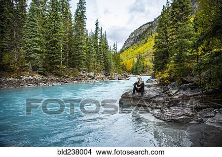 Picture - Caucasian woman sitting on rock near mountain river. Fotosearch