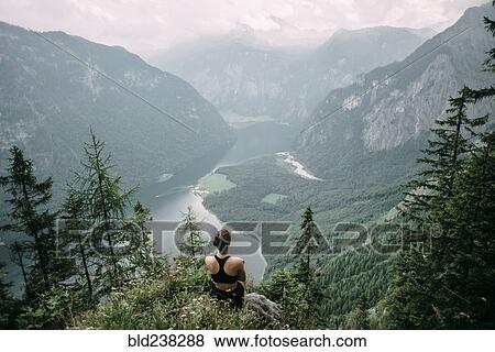 Stock Photo - Caucasian woman sitting on rock overlooking lake in valley. Fotosearch