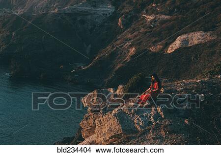 Picture - Caucasian woman sitting on rocks at ocean. Fotosearch