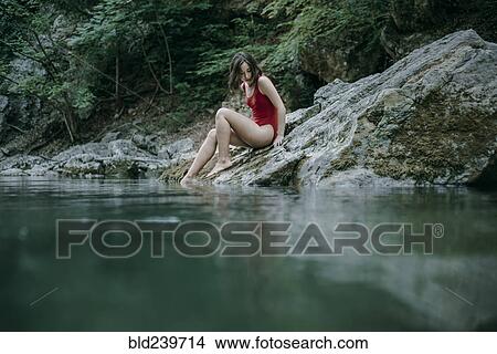 Picture - Caucasian woman sitting on rocks at pool of water. Fotosearch
