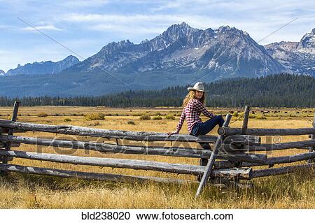 Stock Image - Caucasian woman sitting on wooden fence near mountain. Fotosearch