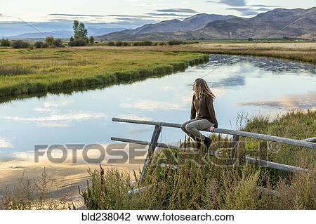 Stock Image - Caucasian woman sitting on wooden fence near mountain river. Fotosearch