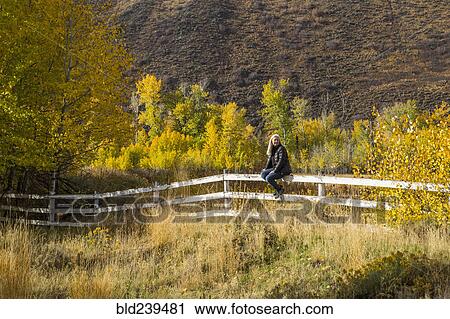 Stock Image - Caucasian woman sitting on wooden fence. Fotosearch
