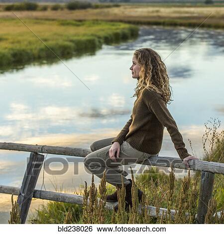 Stock Photograph - Caucasian woman sitting on wooden fence near river. Fotosearch