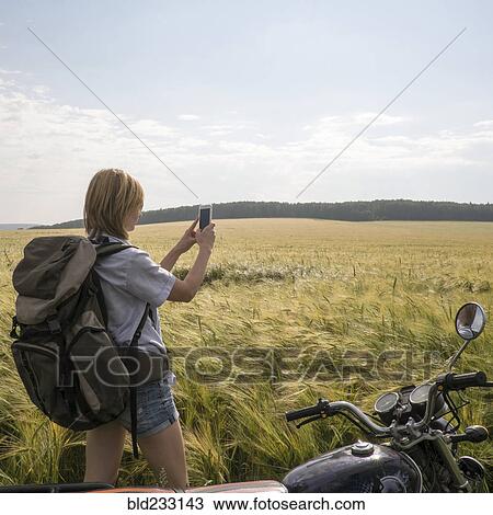 Stock Image - Caucasian woman standing in field near motorcycle using cell phone. Fotosearch