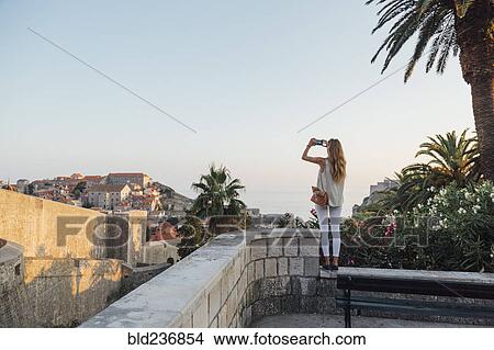 Caucasian woman standing on park wall photographing cityscape View Large Photo Image Picture - Caucasian woman standing on park wall photographing cityscape. Fotosearch