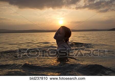Stock Image - Caucasian woman swimming in lake at sunset. Fotosearch