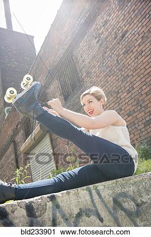 Stock Image - Caucasian woman tying roller skate near brick wall. Fotosearch