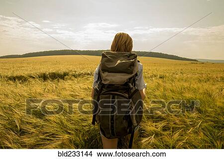 Picture - Caucasian woman walking in field carrying backpack. Fotosearch