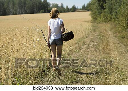 Stock Image - Caucasian woman walking in field carrying basket and stick. Fotosearch