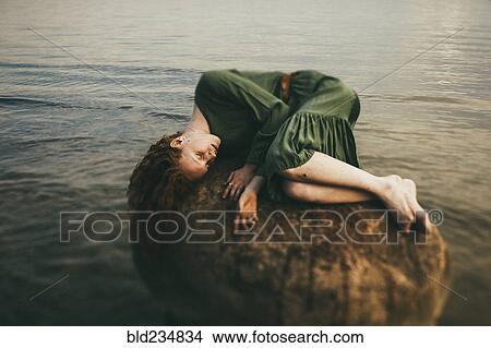 Picture - Caucasian woman wearing dress laying on rock at beach. Fotosearch